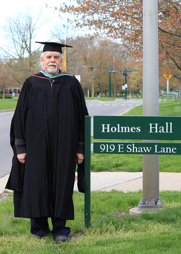 Kent Workman in academic regalia by the sign for Holmes Hall