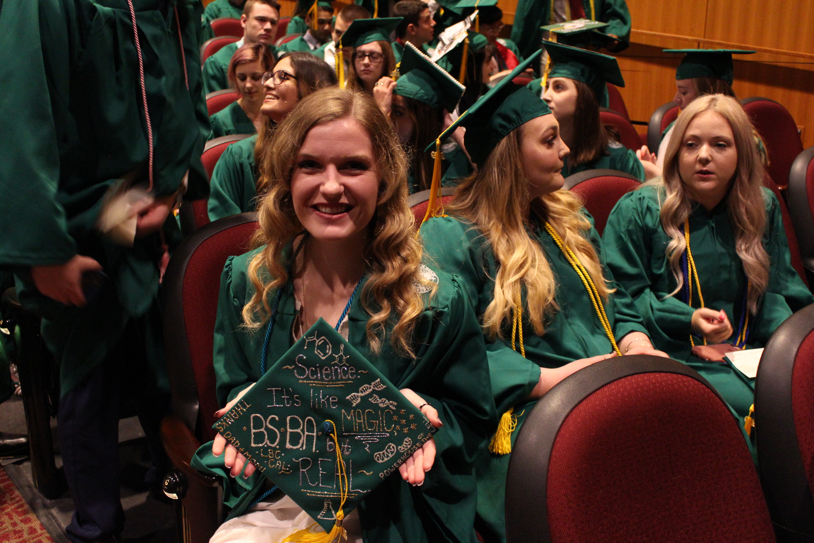 On graduation day, students decorated their mortarboard hats. One says "Science is like magic but real" in glittery writing with cellular structures and glittery writing