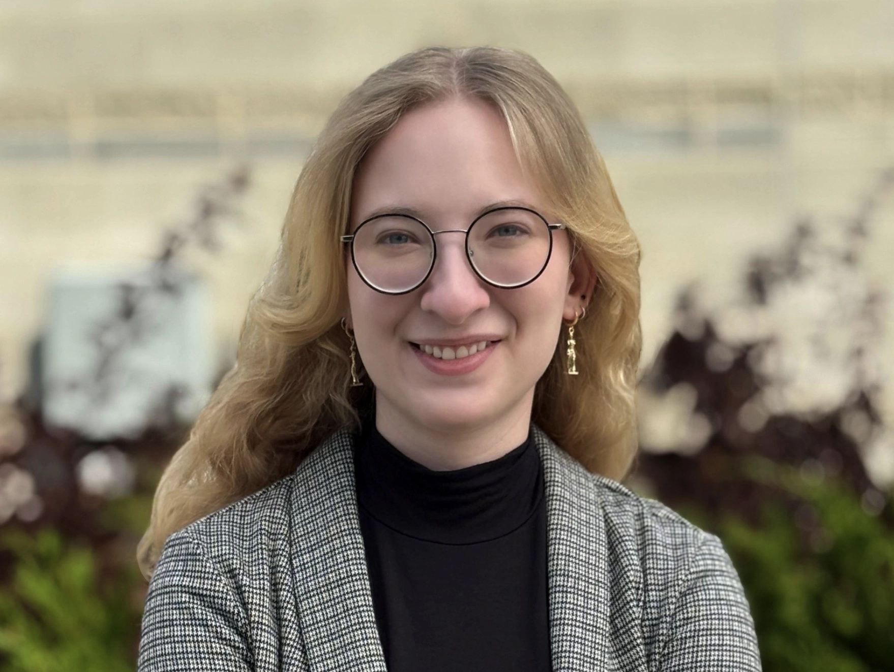 Kierra Jursch has blond wavy hair and glasses. She is smiling and is wearing a checkered blazer and black turtleneck top
