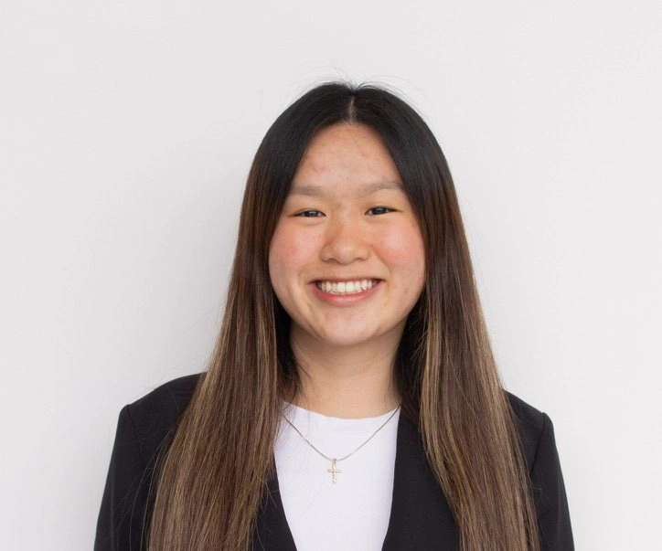 Tammy Thai smiles. She is wearing a black blazer and white top, with a cross necklace. Her hair is brown and long.