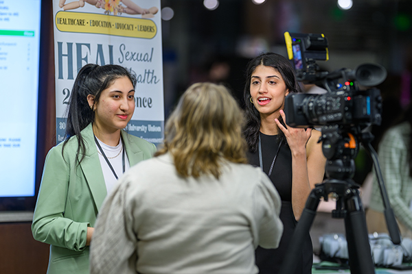 Nupur Huria and Harsna Chahal talk to a reporter while standing next to the HEAL conference sign