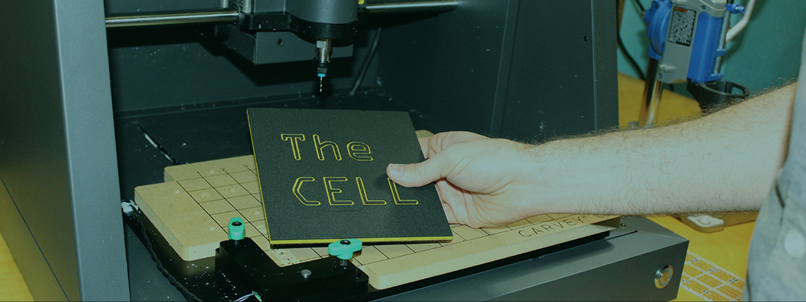 A hand holds a laser-engraved piece of acetate with the words The CELL