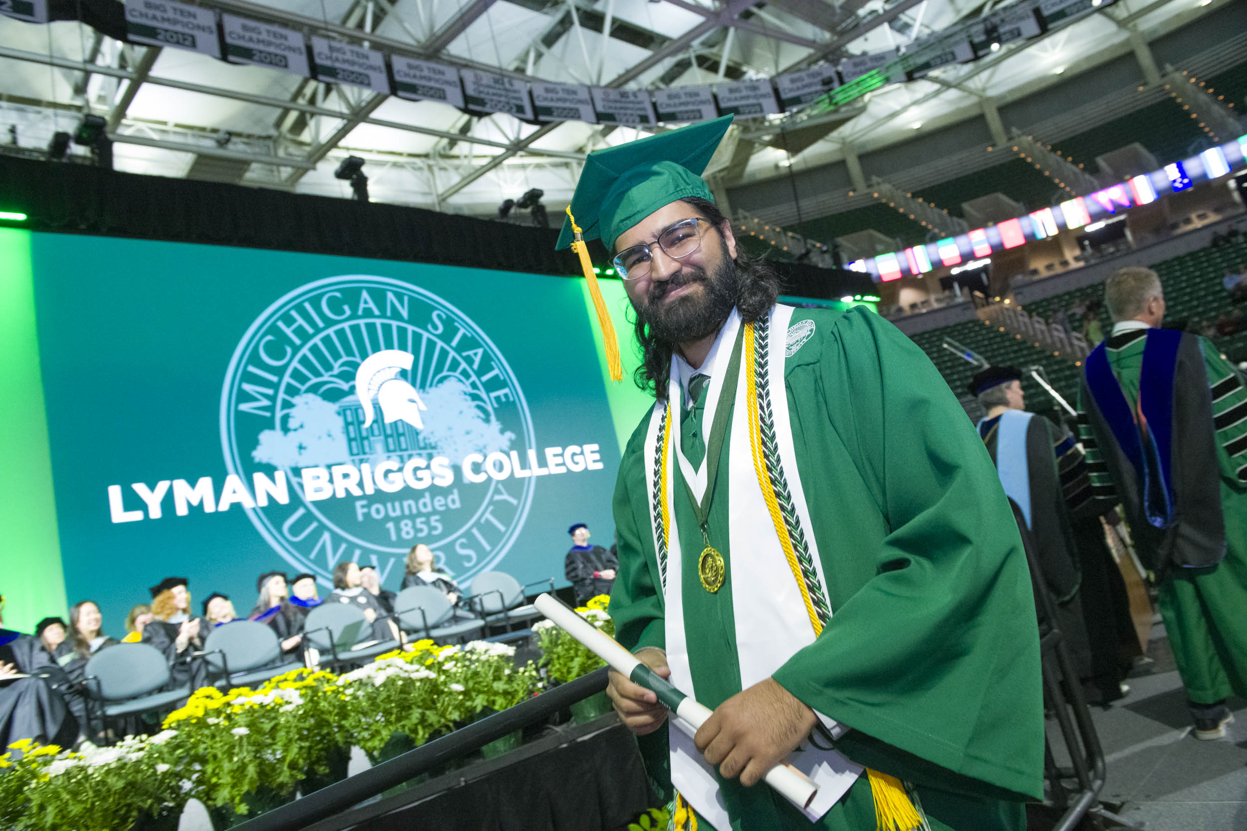 A Spring 2025 LBC graduate smiles after receiving their diploma. Bright green Lyman Briggs College screen shows behind