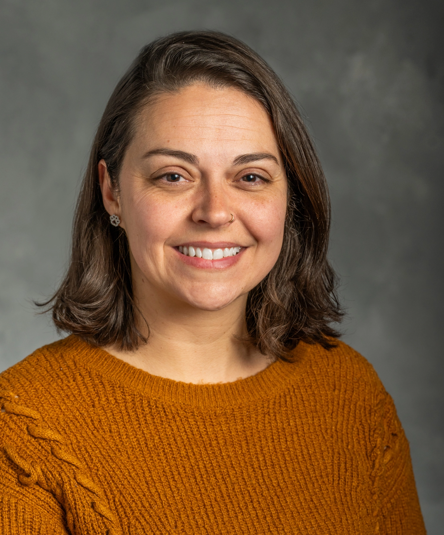 Amanda Domingues has shoulder-length brown hair, and is wearing a muted orange sweater in this portrait