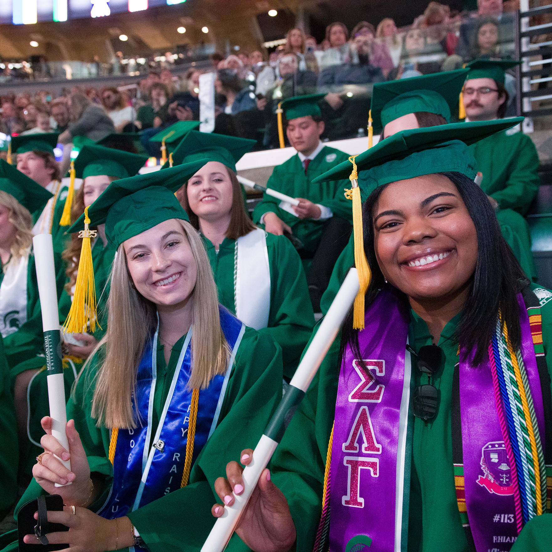 Two graduates with diplomas in hand smile at the 2025 spring commencement