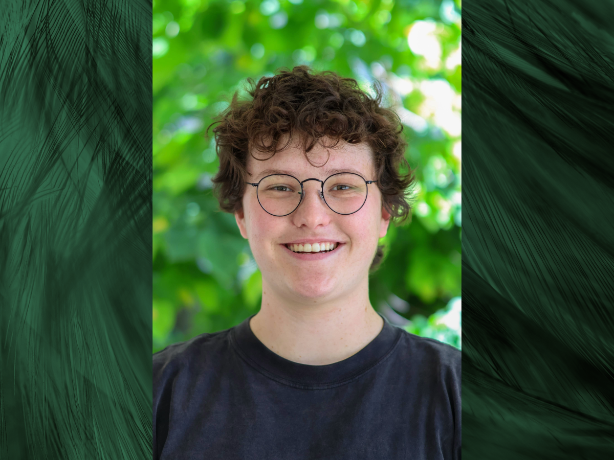 Emma Andrechek has short curly brown hair and wears glasses and a black top. Emma is smiling and there are green trees in the background
