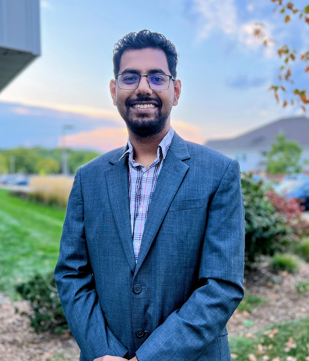 Maruf wears a suit jacket and a plaid dress shirt. He is smiling. He has short black hair, facial hair, and wears glasses.