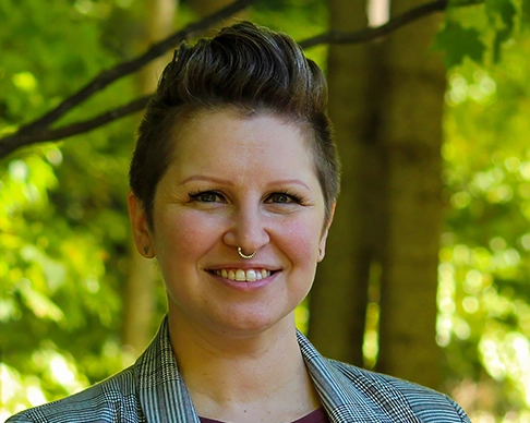 Andrea Kelley has short hair that is longer on top. They are smiling, are wearing a plaid jacket, and have a small nose ring. 