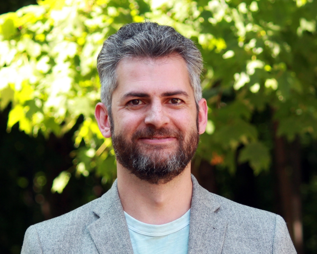 Isaac Record is smiling. He has greying hair, a dark beard and moustache, and wears a pale green shirt with a gray blazer. 