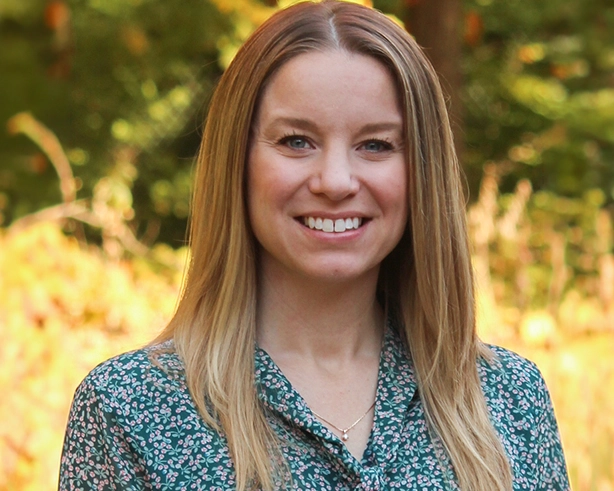 Kristen Vroom has medium-length blonde hair, and wears a flowered shirt. The background is yellow 