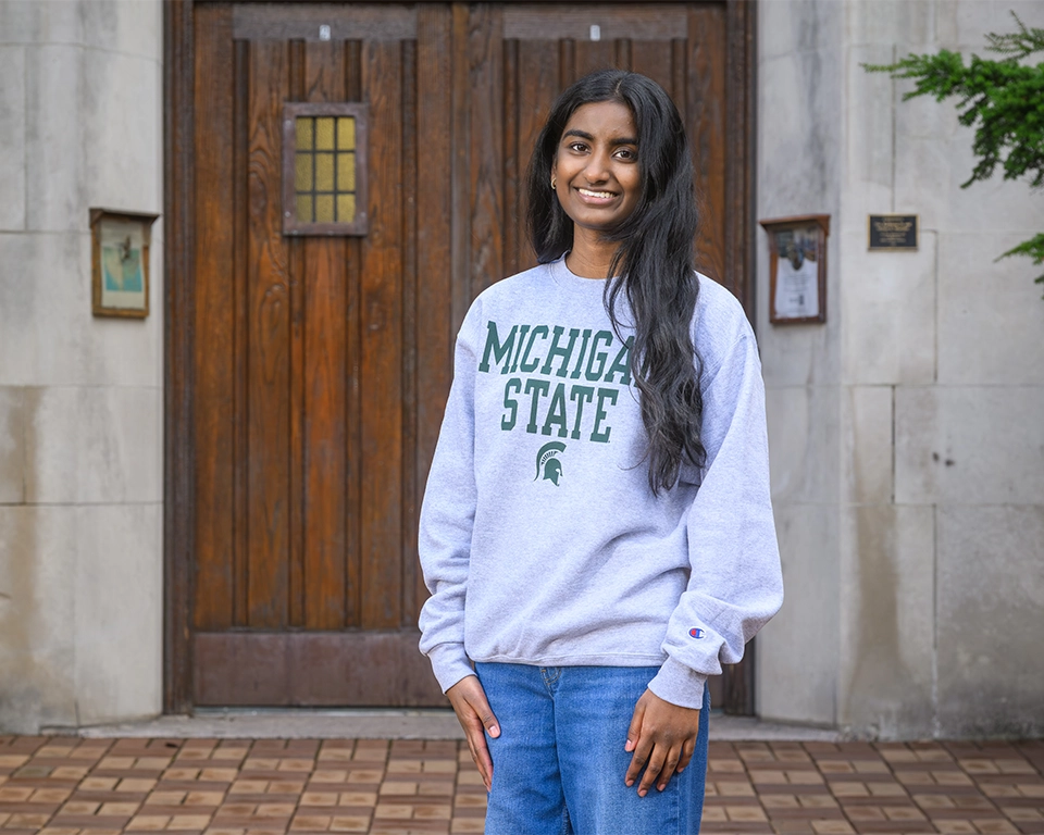 Shrinidhi Pola is standing at the base of Beaumont Tower. She wears a white Michigan State crewneck sweatshirt and medium blue jeans. She has long, dark hair 