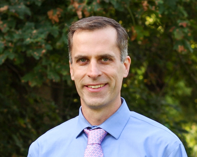 Ryan Sweeder wears a blue dress shirt and pale pink tie. He has dark hair that is greying on the sides