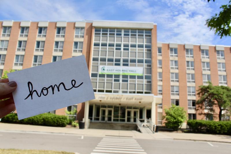 Front of Holmes Hall with someone holding a card in front that says home