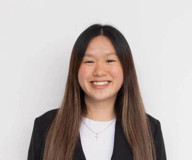 Tammy Thai has long brown hair and is smiling. She is wearing a black jacket, a white top, and a cross necklace