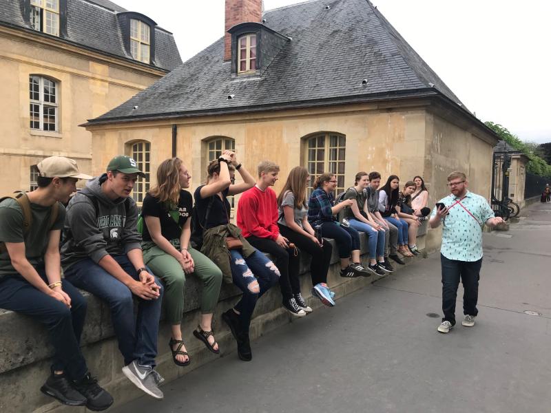 Students sit along a wall at a chateau in France as another student presents