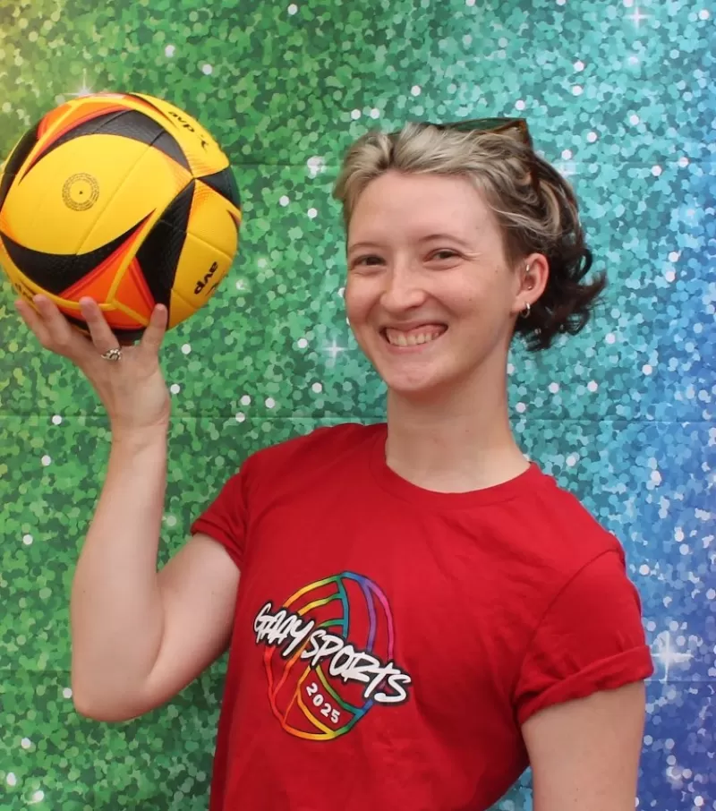 C Davis wears a red shirt with a volleyball on it outlined in rainbow colors. They are smiling and are holding a yellow volleyball