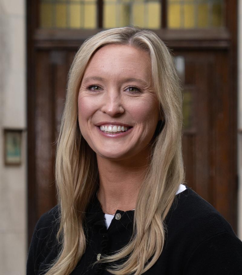 Jessie Pung stands by Beaumont Tower. She has medium-length blond hair and is smiling. She is wearing a white shirt and a black buttoned shirt over it. 