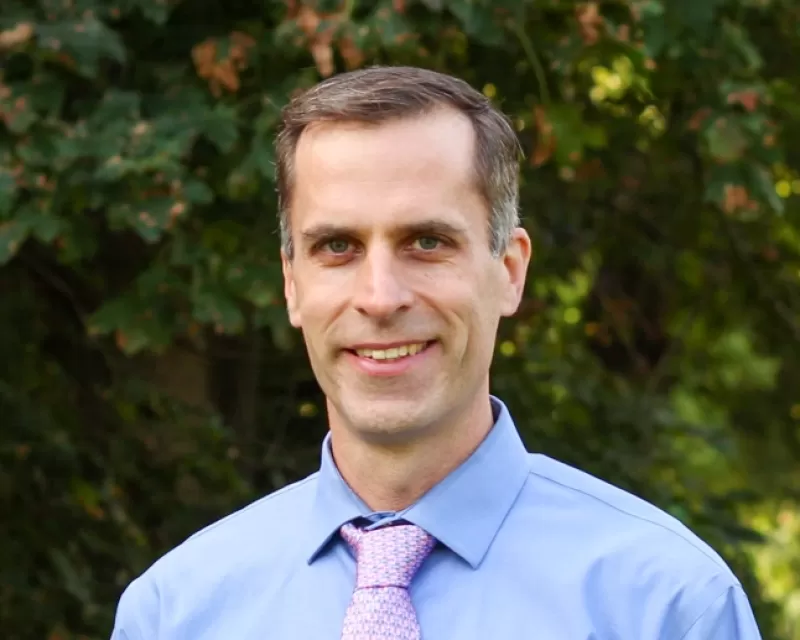 Ryan Sweeder wears a blue dress shirt and pale pink tie. He has dark hair that is greying on the sides